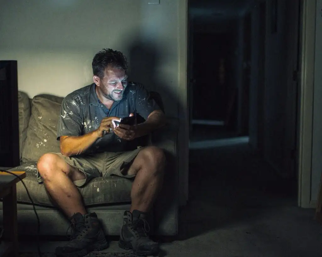 The same tradesman on the same couch in the same dark lounge room, sitting forward with the phone in both hands, replying, half-smiling.