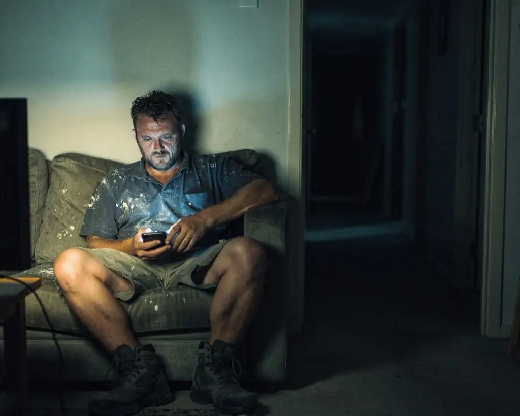 A tradesman still in paint-spattered work clothes and boots, slumped into the corner of a couch in a dark lounge room at night, face lit by the phone in his hands.