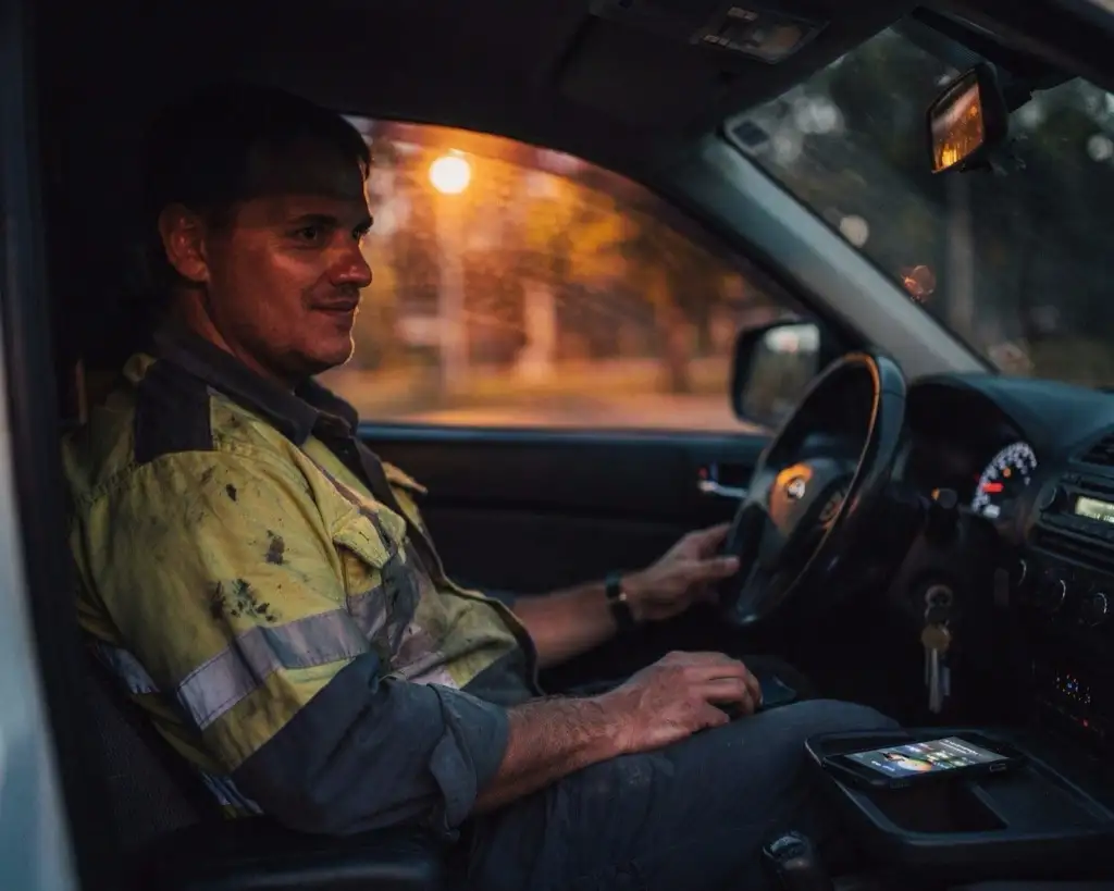 The same tradesman in the same ute at dusk, leaning back with a hand on the wheel and his phone put down beside him, looking out through the windscreen.
