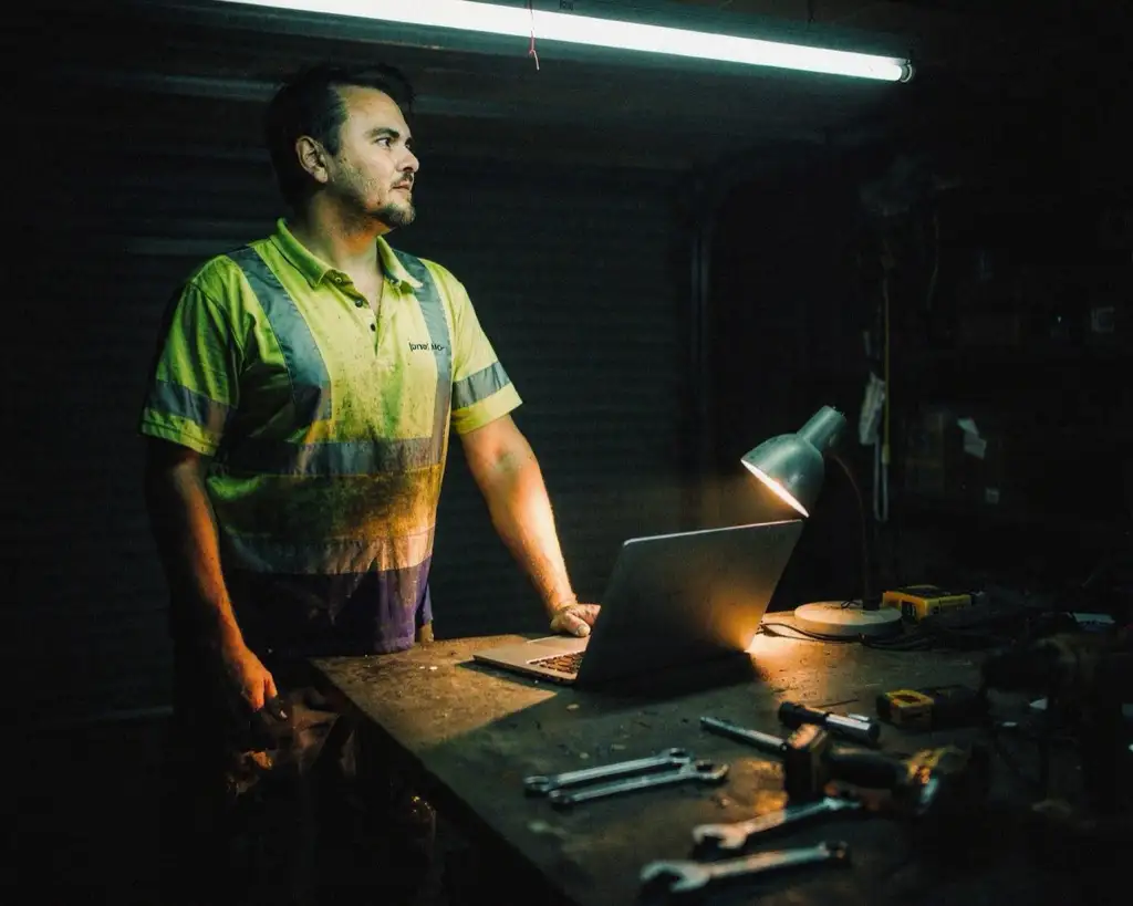 The same tradesman at the same garage workbench, standing straight now with the laptop shut and the bench clear, looking away with the desk lamp still on.